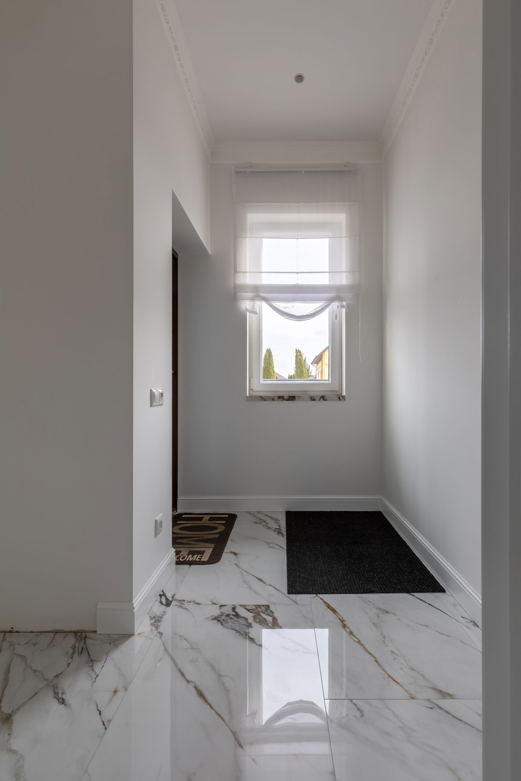 A bright and minimalist entryway featuring marble flooring, white walls, a window with sheer curtains, and a modern aesthetic.