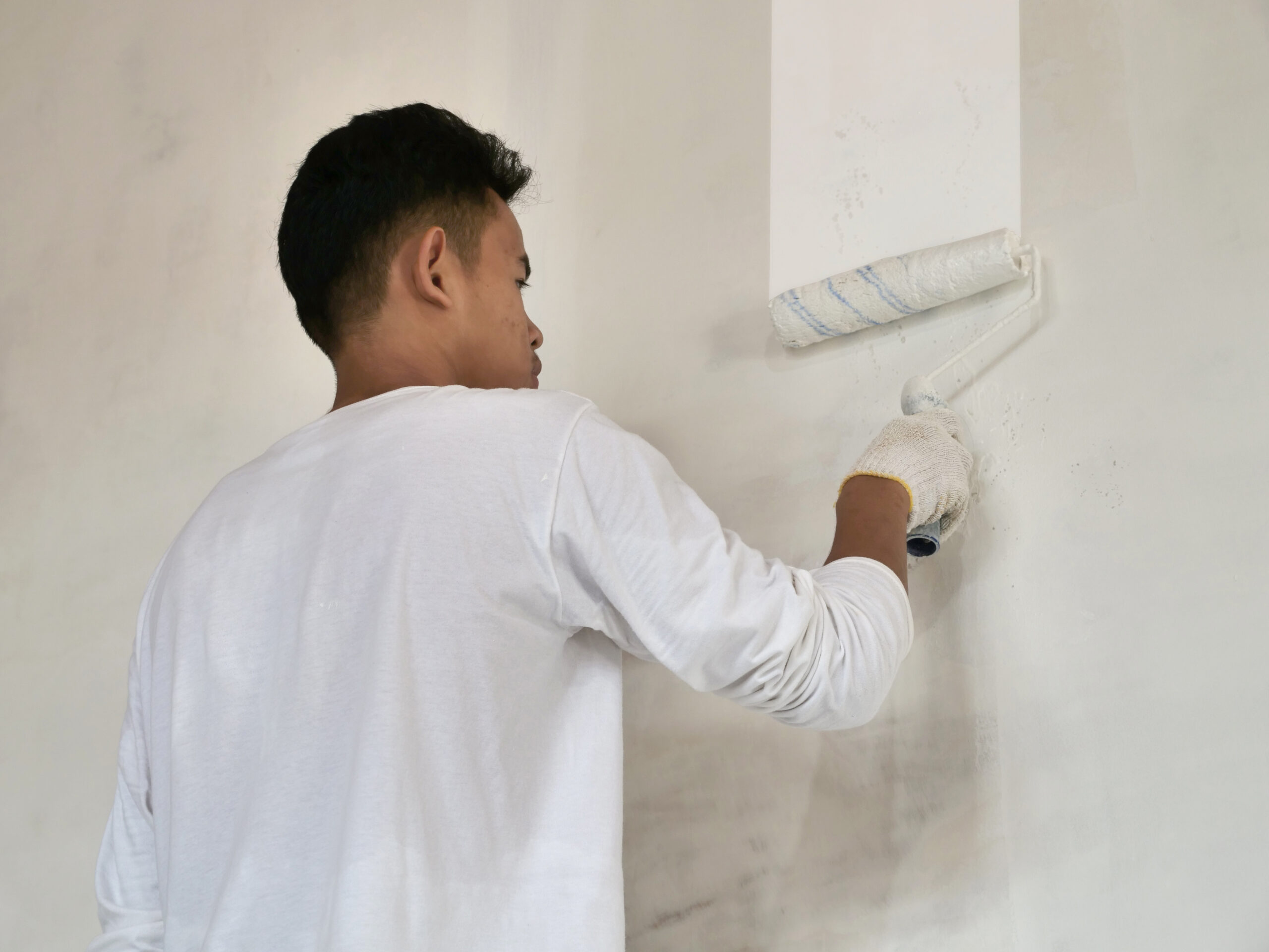 Asian male painter painting new white wall with roller brush, house interior renovation concept