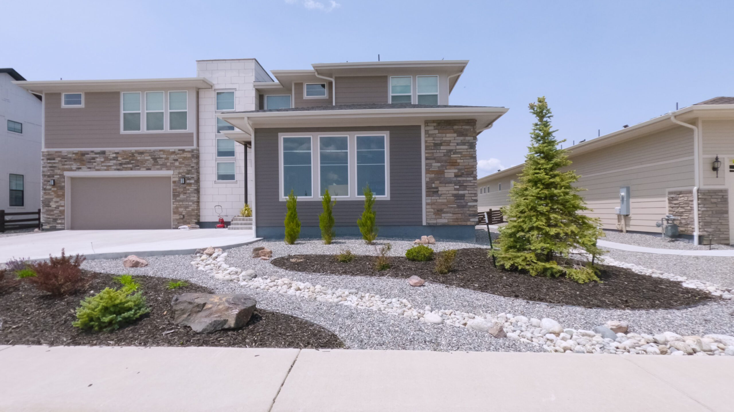 A two-story home with tall front windows, light gray siding, and stone facade. The yard includes a rock bed, young shrubs, and wide driveway.