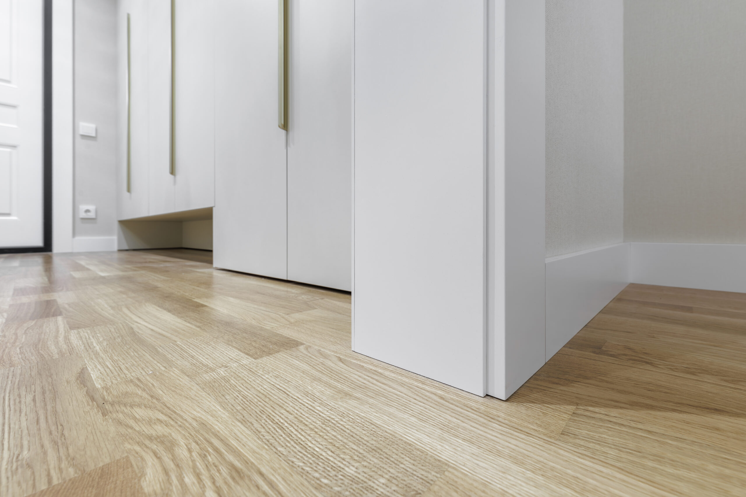 A low-angle architectural shot focusing on the clean transition between a white minimalist wall, a wide skirting board, and natural oak parquet flooring. The background features sleek white built-in cabinetry with elegant long golden handles.